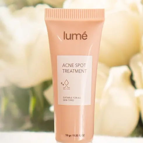 Tube Lume Acne Spot Treatment berdiri di atas kain krem di depan bunga putih