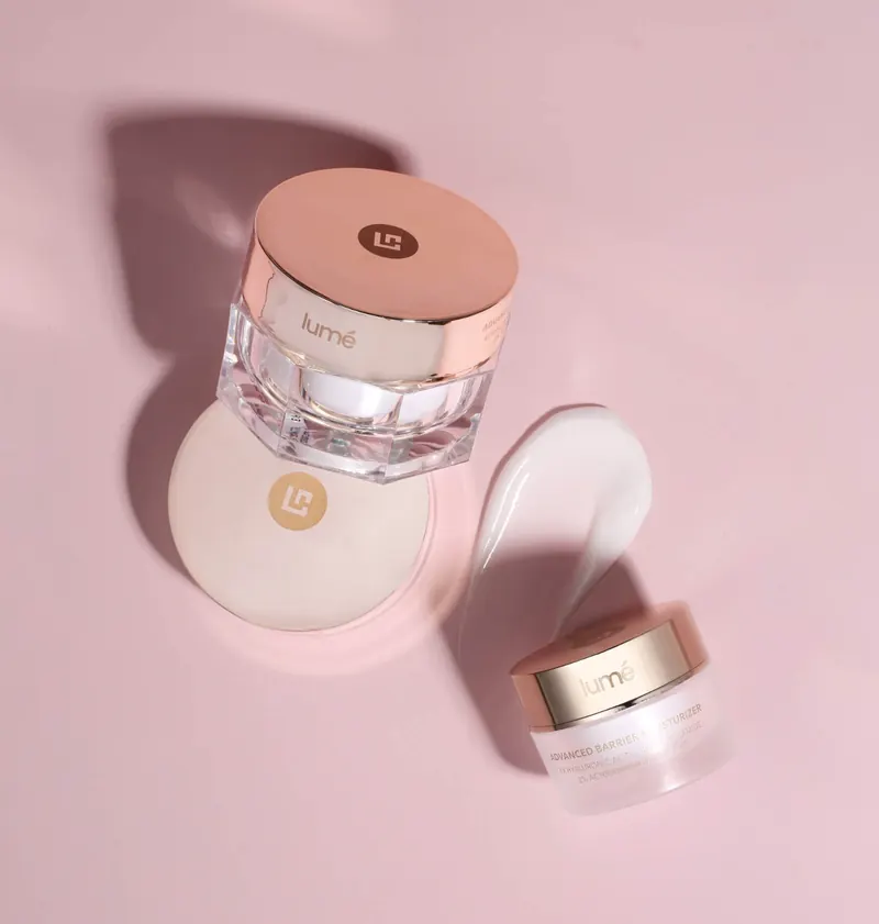 Dua jar Lume Advanced Barrier Moisturizer bertutup rose gold dan olesan krim putih di atas latar merah muda