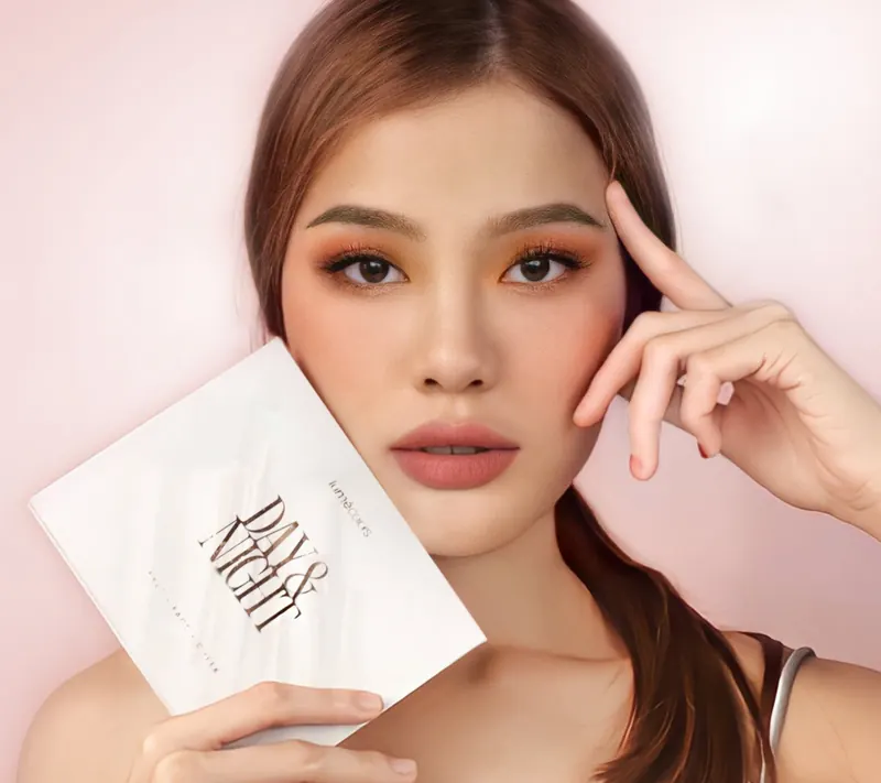 Model berambut cokelat kemerahan dengan eyeshadow peach menempelkan kotak Lumécolors Day & Night ke pipinya