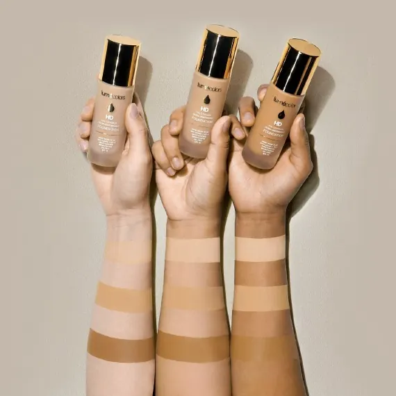 Tiga lengan berkulit terang, medium dan gelap mengangkat botol HD Foundation, dengan olesan swatch di tiap lengan
