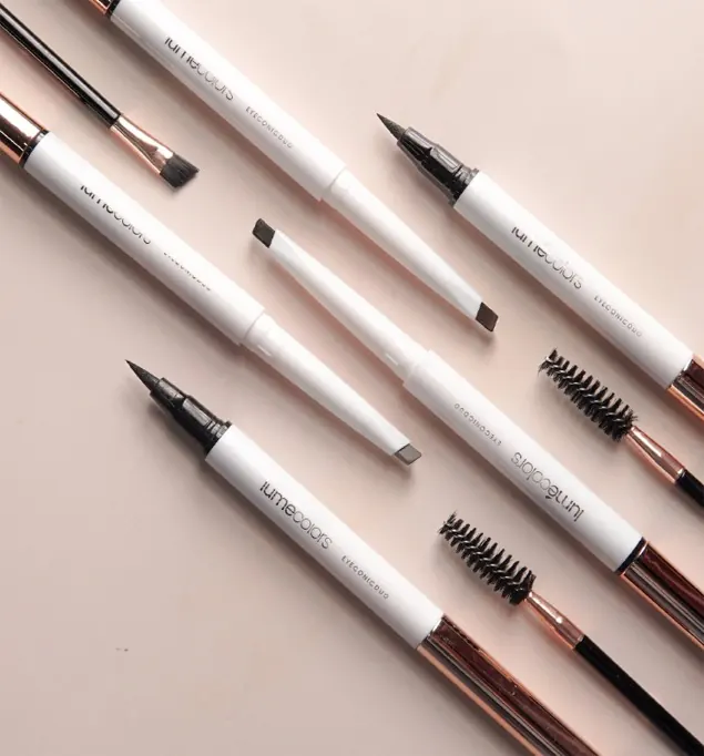 Beberapa pensil Lumécolors Eyeconic Duo dan sikat spoolie tersusun diagonal di atas latar pink