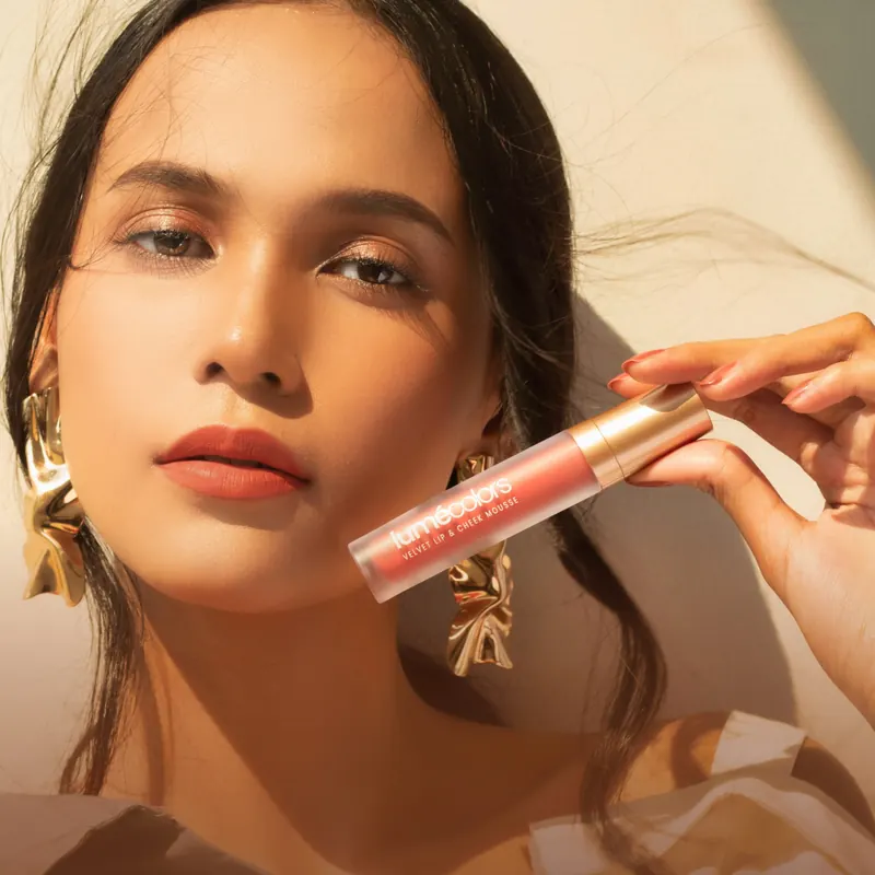 Model berbibir terracotta-nude matte memegang tube Lumécolors Velvet Lip & Cheek Mousse di pipinya