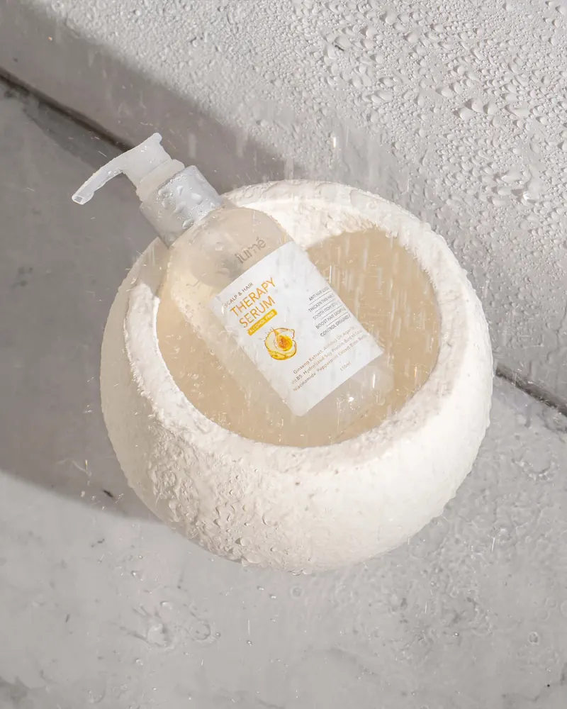 Botol Lume Scalp & Hair Therapy Serum di dalam mangkuk batu putih yang basah di bawah pancuran